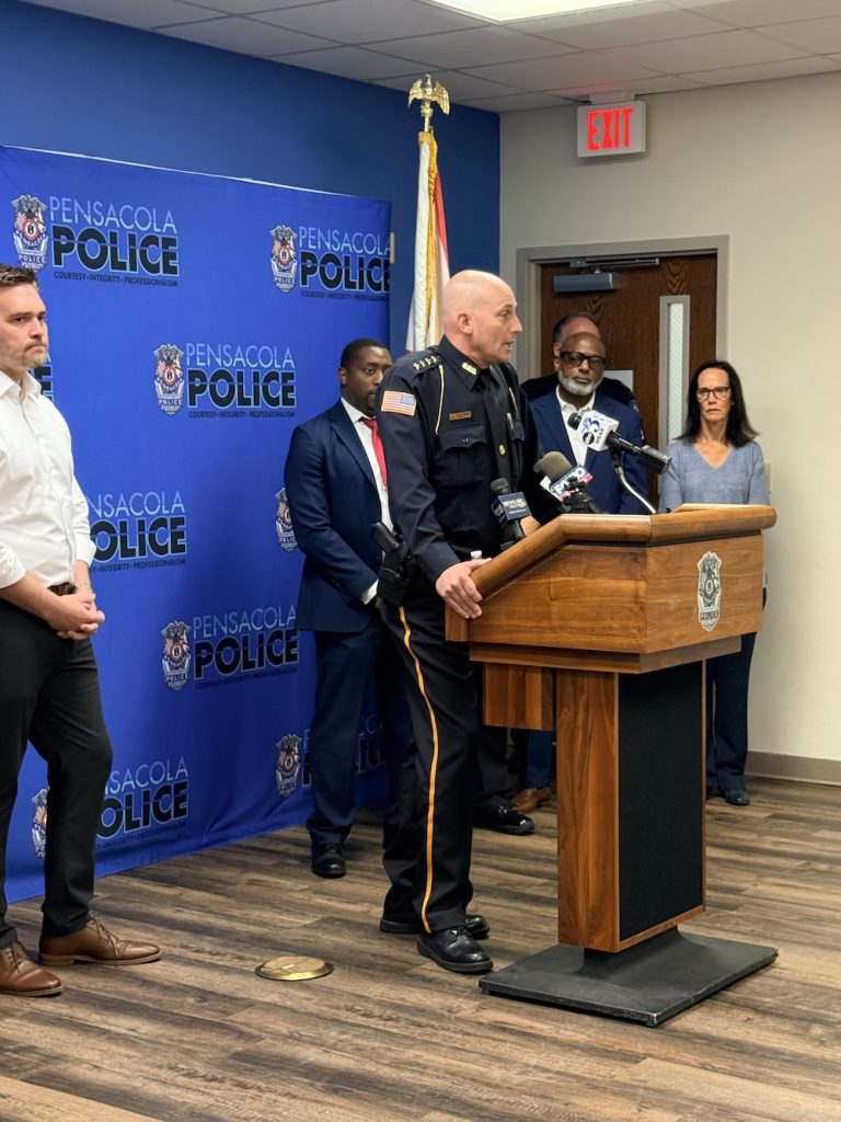 Police chief in dress uniform speaking at a Pensacola Police press conference, with microphones on the wooden podium and officials behind him.”,