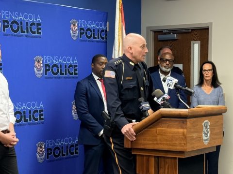 Police chief in dress uniform speaking at a Pensacola Police press conference, with microphones on the wooden podium and officials behind him.”,