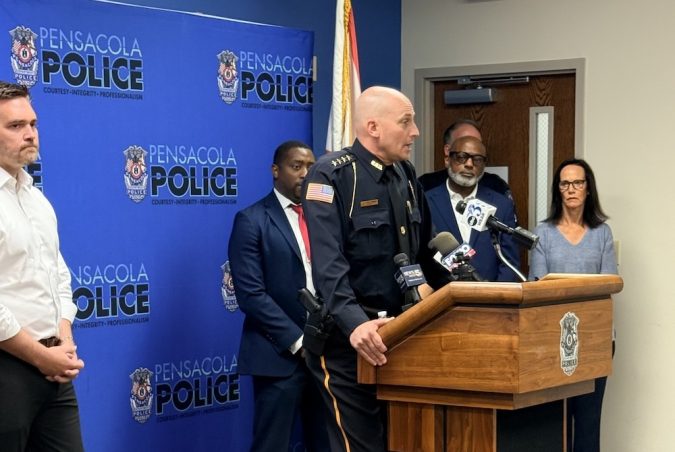 Police chief in dress uniform speaking at a Pensacola Police press conference, with microphones on the wooden podium and officials behind him.”,