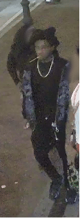 Person walking on a sidewalk wearing a black outfit and a blue patterned jacket, with a pearl necklace and cat-ear headband.