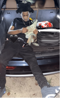 Young man in a cap sitting in the open trunk of an SUV, counting a stack of cash.