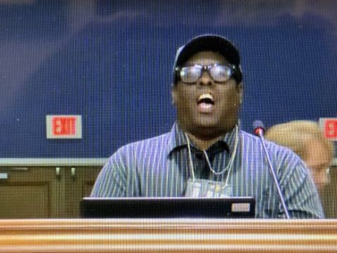 Man wearing a striped shirt and cap speaks into a microphone at a podium in a conference room.