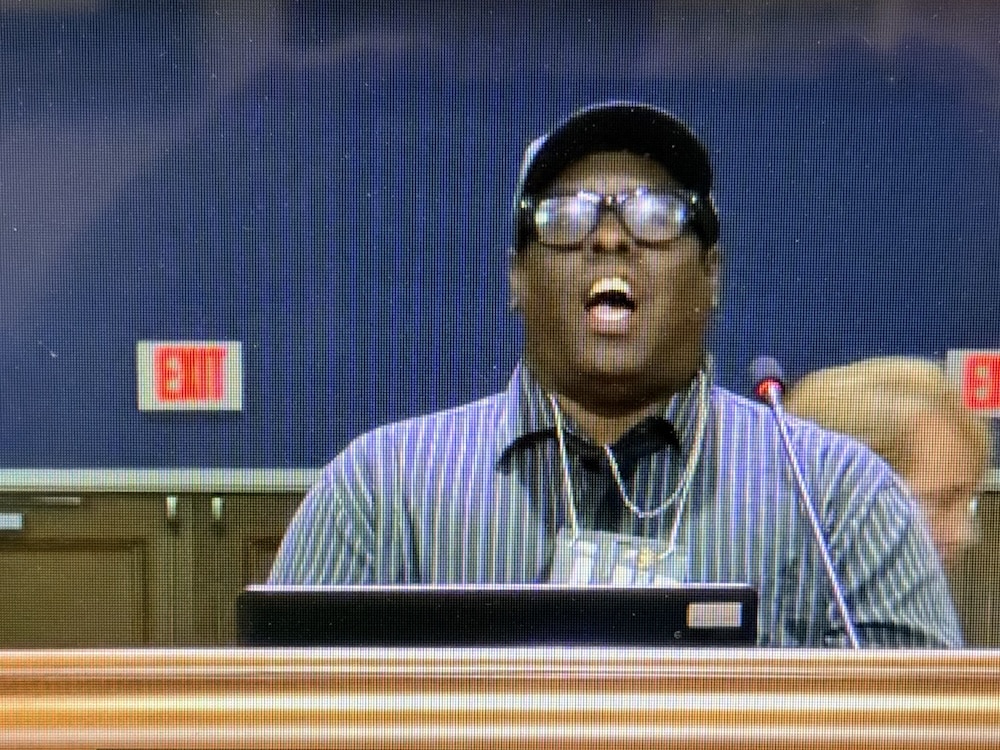 Man wearing a striped shirt and cap speaks into a microphone at a podium in a conference room.