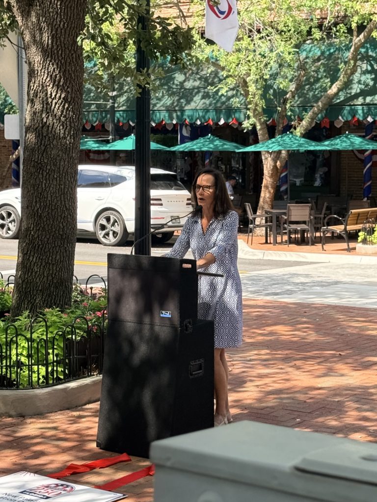 Woman in a geometric dress speaks at a podium outdoors beside a tree-lined street with cars and cafe seating behind her.