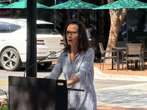 Woman in a geometric dress speaks at a podium outdoors beside a tree-lined street with cars and cafe seating behind her.