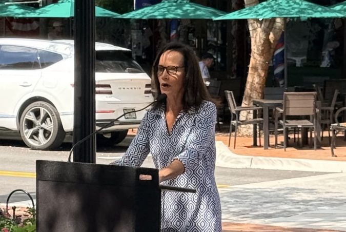 Woman in a geometric dress speaks at a podium outdoors beside a tree-lined street with cars and cafe seating behind her.