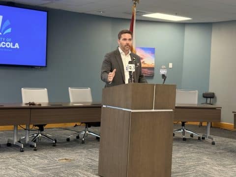 Man in a suit speaks at a wooden podium with microphones in a conference room.