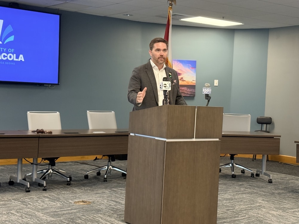 Man in a suit speaks at a wooden podium with microphones in a conference room.