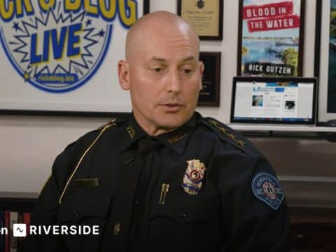 Bald police officer in full uniform seated in an office, with framed posters and a laptop on a desk behind him.