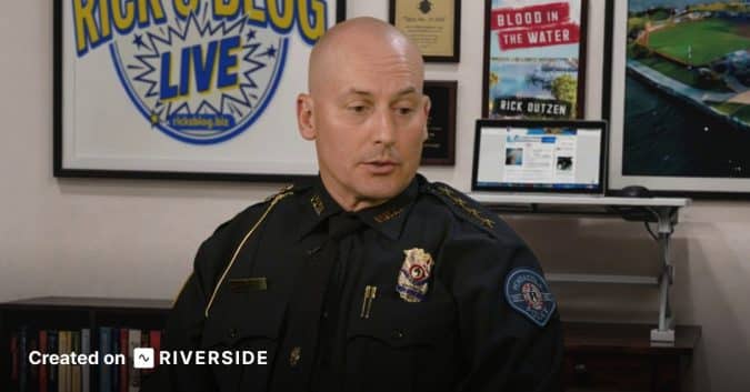 Bald police officer in full uniform seated in an office, with framed posters and a laptop on a desk behind him.