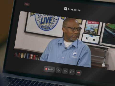 Man in a blue shirt and glasses on a video call, studio backdrop with posters and a Riverside logo behind him