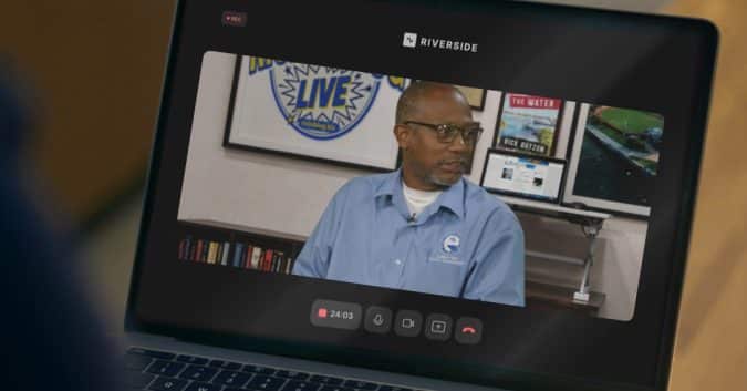 Man in a blue shirt and glasses on a video call, studio backdrop with posters and a Riverside logo behind him