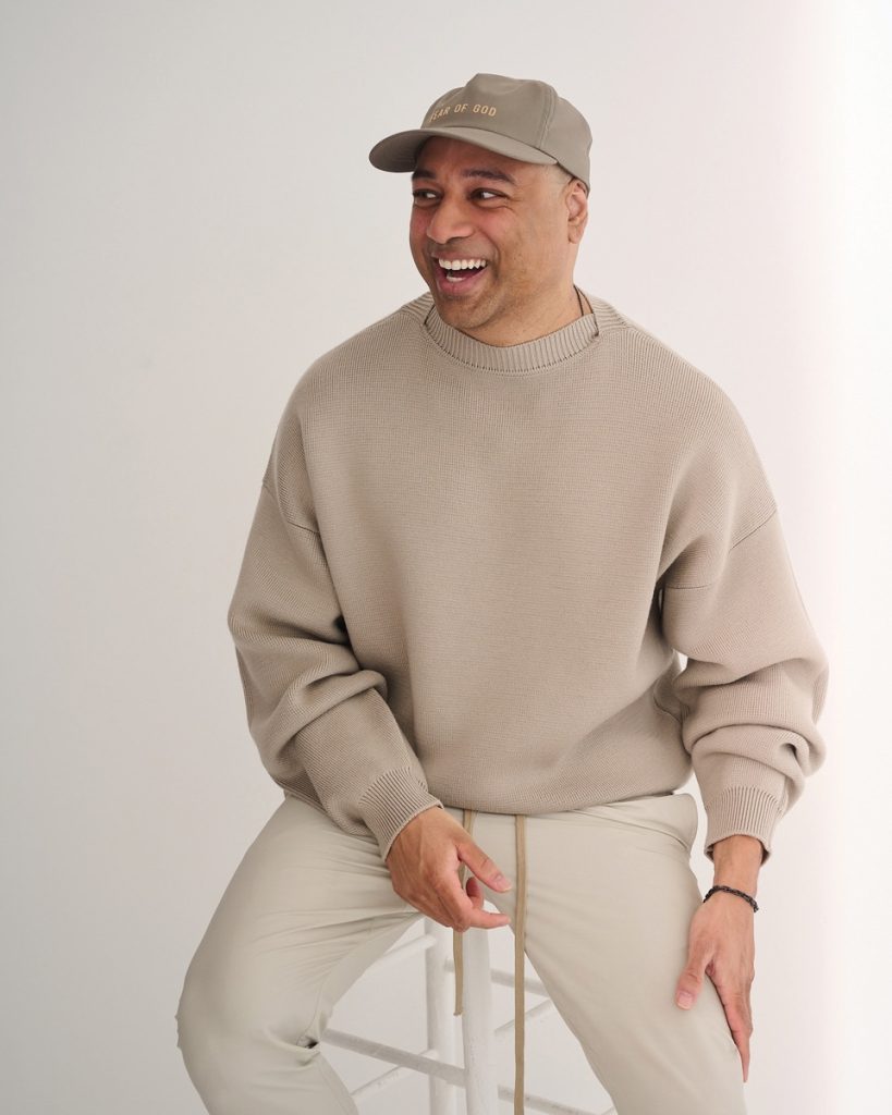 Man smiling and looking to his left, wearing a beige knit sweater and a tan cap against a light background.