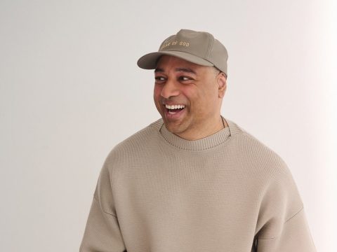 Man smiling and looking to his left, wearing a beige knit sweater and a tan cap against a light background.
