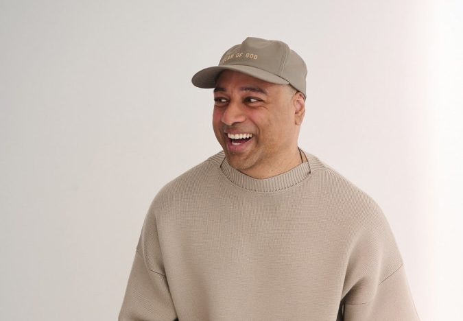 Man smiling and looking to his left, wearing a beige knit sweater and a tan cap against a light background.