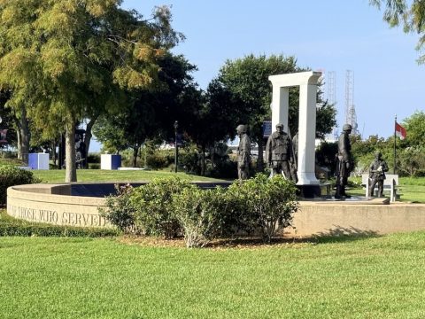 War memorial with a circular stone basin and a row of metal soldier statues in a park. A white arch monument stands behind them.