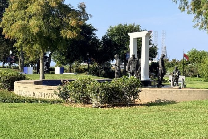 War memorial with a circular stone basin and a row of metal soldier statues in a park. A white arch monument stands behind them.