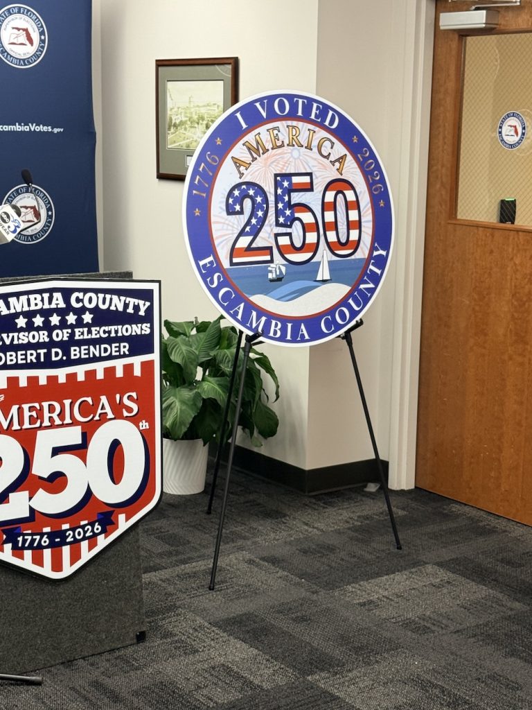 Circular 'I Voted America 250' sign for Escambia County, Florida, displayed on a metal easel in an office setting with other election signage nearby.