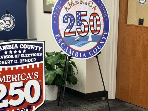 Circular 'I Voted America 250' sign for Escambia County, Florida, displayed on a metal easel in an office setting with other election signage nearby.
