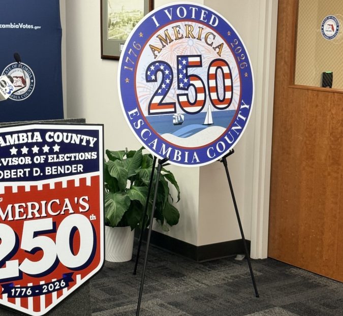Circular 'I Voted America 250' sign for Escambia County, Florida, displayed on a metal easel in an office setting with other election signage nearby.