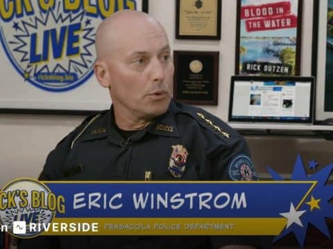 Bald Pensacola Police Department officer sits in a studio interview, with a blue-yellow banner reading 'Eric Winstrom' and 'Rick's Blog Live'.