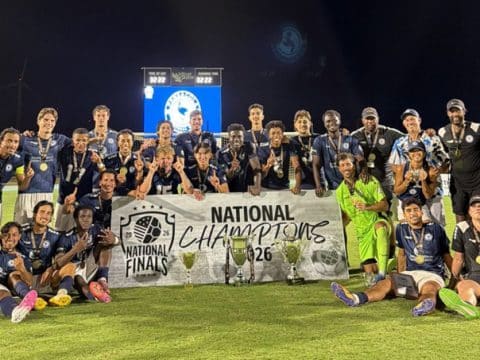 Soccer team posing on a field behind a banner that reads 'National Champions' with trophies and medals at night stadium lights.