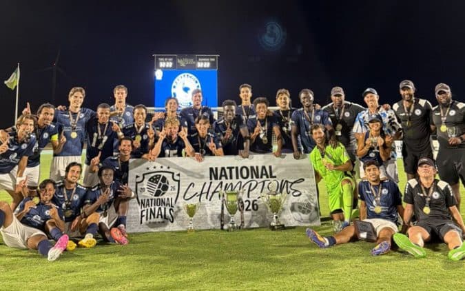 Soccer team posing on a field behind a banner that reads 'National Champions' with trophies and medals at night stadium lights.