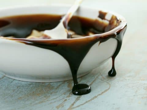 White ceramic bowl with melted dark chocolate dripping over the rim and onto a pale surface, spoon inside partially visible.