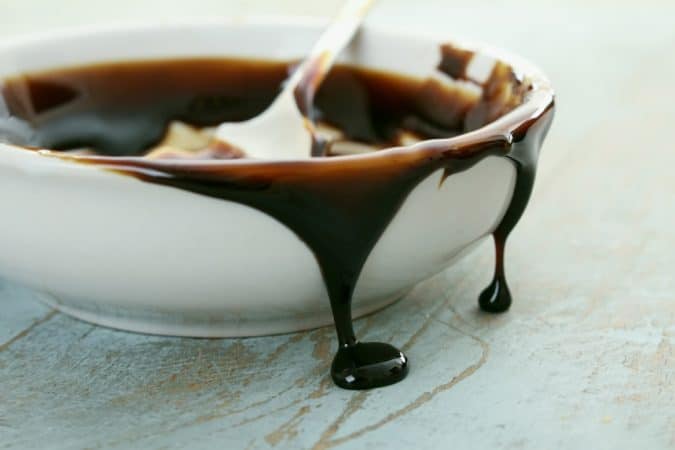 White ceramic bowl with melted dark chocolate dripping over the rim and onto a pale surface, spoon inside partially visible.