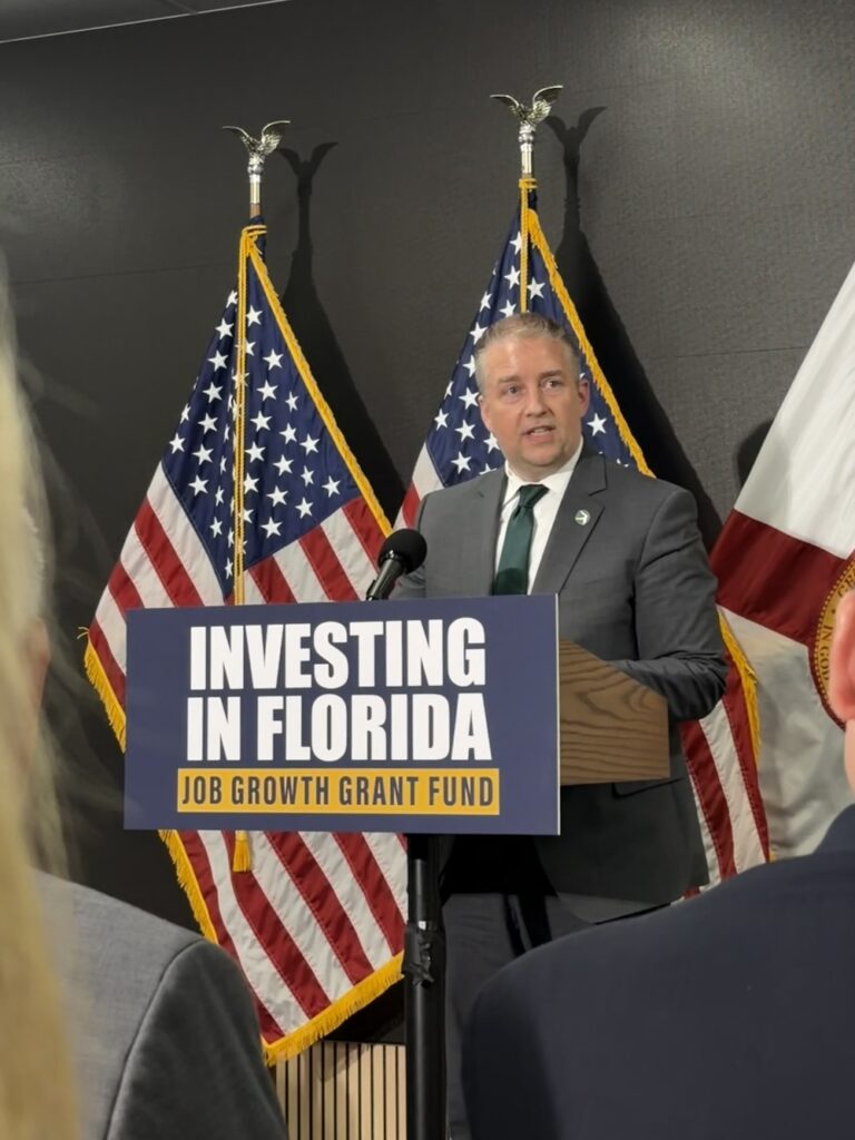 Man in a suit giving a speech at a podium with a sign reading 'Investing in Florida, Job Growth Grant Fund' behind him are U.S. flags and a state flag,