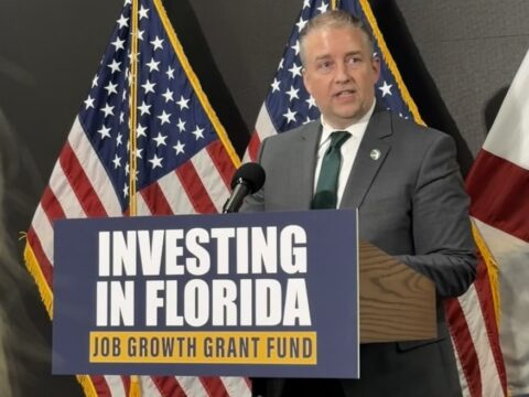 Man in a suit giving a speech at a podium with a sign reading 'Investing in Florida, Job Growth Grant Fund' behind him are U.S. flags and a state flag,