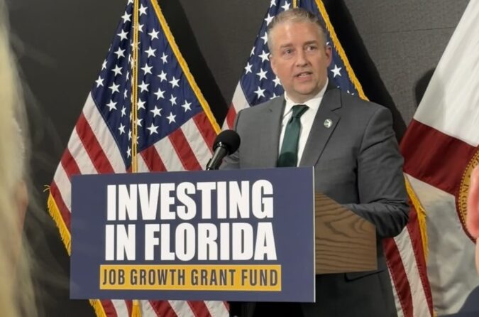 Man in a suit giving a speech at a podium with a sign reading 'Investing in Florida, Job Growth Grant Fund' behind him are U.S. flags and a state flag,