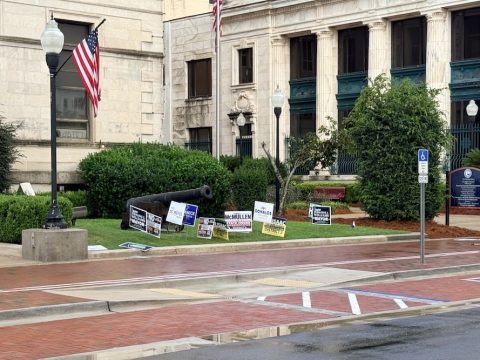 A black cannon on a grassy patch in front of a stone government-style building, with campaign signs, flags, and lampposts nearby.