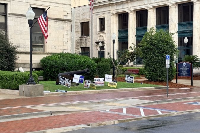 A black cannon on a grassy patch in front of a stone government-style building, with campaign signs, flags, and lampposts nearby.