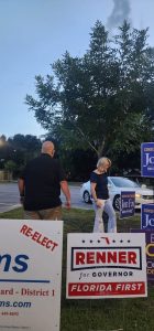 A bald man in a black shirt and a woman with blonde hair in a dark blue top walk past campaign signs in a parking-lot grass area; signs say 'RENNER for Governor' and 'Florida First'.