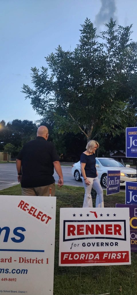 A bald man in a black shirt and a woman with blonde hair in a dark blue top walk past campaign signs in a parking-lot grass area; signs say 'RENNER for Governor' and 'Florida First'.