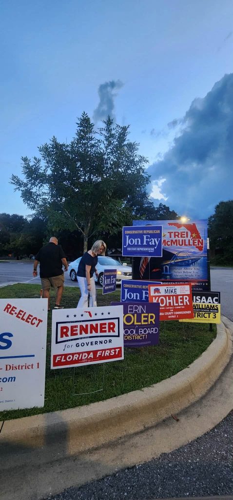Campaign signs for Jon Fay and other candidates are displayed along a grassy curb, with a person bending down near signs by a blue car at dusk.