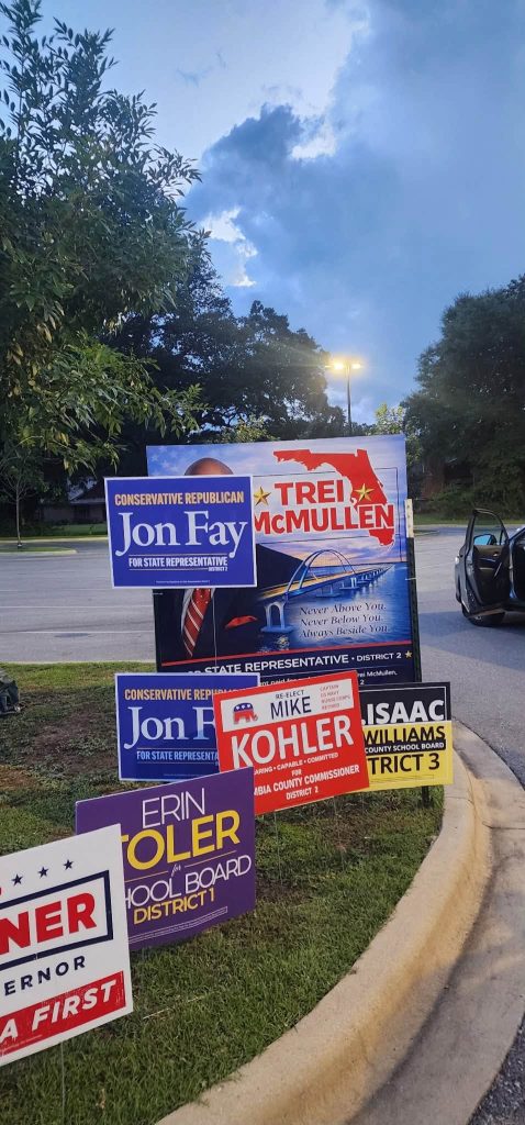 Campaign yard signs along a curb featuring multiple candidates, including Jon Fay for State Representative and Tre McMullen, with a large blue sign and a smaller white-and-blue sign in the foreground.