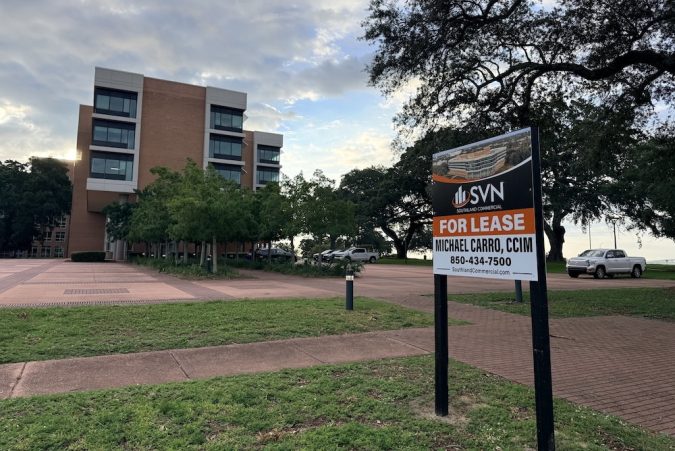 Commercial building with a 'For Lease' sign in the foreground near a sidewalk and trees by the parking lot.
