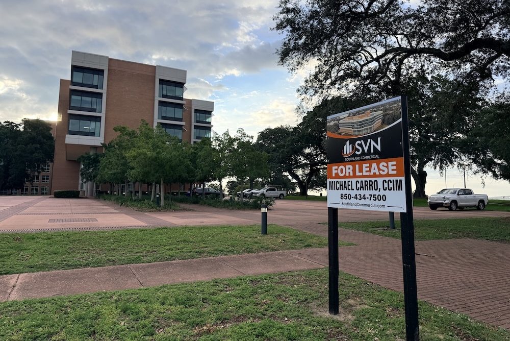 Commercial building with a 'For Lease' sign in the foreground near a sidewalk and trees by the parking lot.
