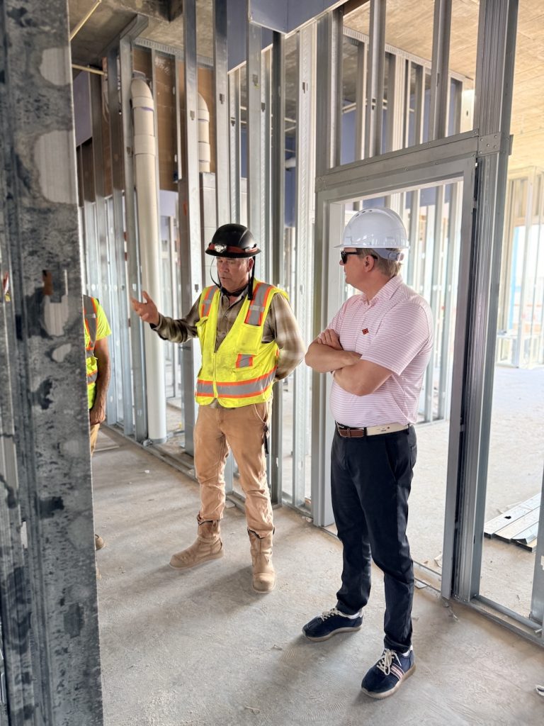 Two construction workers with hard hats and reflective vests discuss inside an unfinished building frame with metal studs and piping visible behind them.