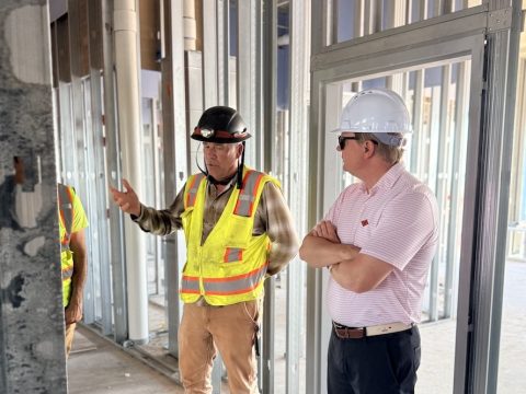 Two construction workers with hard hats and reflective vests discuss inside an unfinished building frame with metal studs and piping visible behind them.