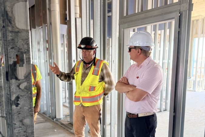 Two construction workers with hard hats and reflective vests discuss inside an unfinished building frame with metal studs and piping visible behind them.