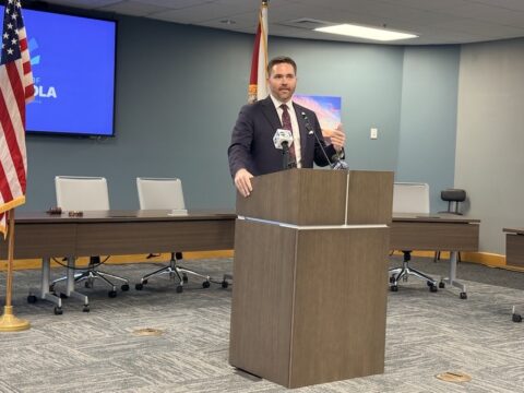 Man in a dark suit speaking at a wooden podium in a conference room, with an American flag and a blue presentation screen behind him.