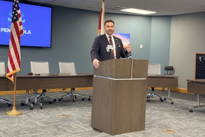 Man in a dark suit speaking at a wooden podium in a conference room, with an American flag and a blue presentation screen behind him.