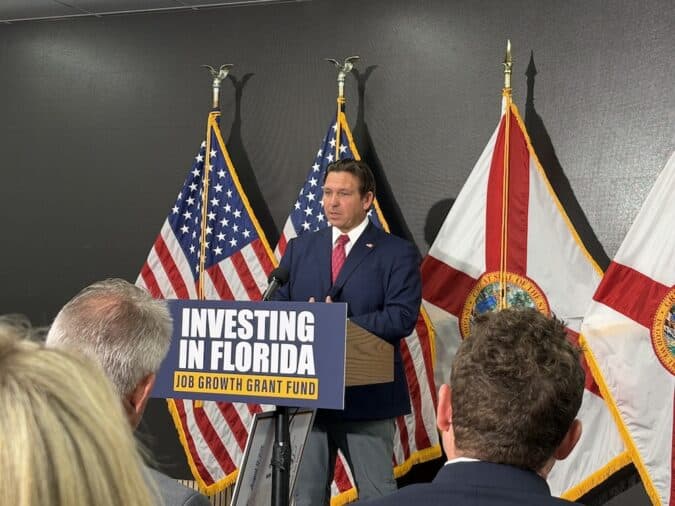 Speaker at a lectern reading 'Investing in Florida' while addressing an audience, with American and state flags in the background.