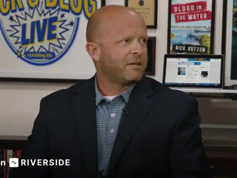 Man in a dark blazer being interviewed in a studio, with framed posters and a laptop in the background.