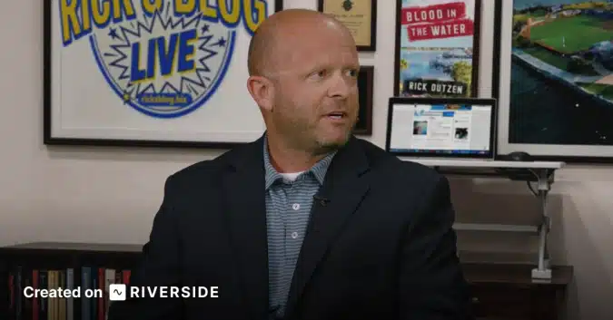 Man in a dark blazer being interviewed in a studio, with framed posters and a laptop in the background.