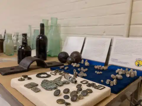 Display of historical artifacts on a table with glass bottles, metal tools, and coins in a museum exhibit setting.