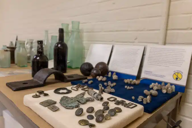Display of historical artifacts on a table with glass bottles, metal tools, and coins in a museum exhibit setting.